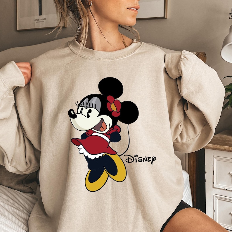 May include: A beige sweatshirt with a cartoon image of Minnie Mouse wearing a red dress and a black bow. The text "DISNEY" is printed below the image.