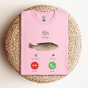 May include: A light pink t-shirt with the words "fih calling..." and a fish graphic. Below the fish are phone icons with the words "Remind Me", "Message", "Decline", and "Accept".