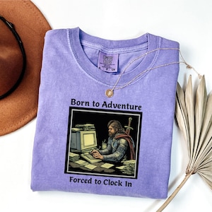 May include: A light purple t-shirt with the text "Born to Adventure, Forced to Clock In" above and below a graphic of a person at a computer. A brown hat and gold necklace are also in the image.
