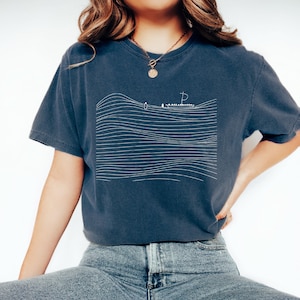 May include: A navy blue t-shirt featuring a minimalist white line drawing of a sailboat on wavy lines. The shirt has short sleeves and a relaxed fit. The design evokes a nautical theme.