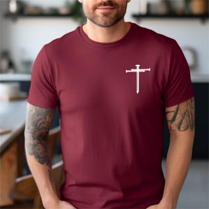 May include: A maroon t-shirt with a white graphic of a cross with nails. The cross is centered on the chest of the shirt.