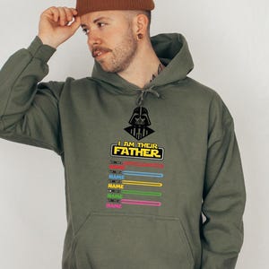 Pu&ograve; includere: Felpa con cappuccio verde oliva con grafica di Darth Vader e la scritta "I AM THEIR FATHER". Sotto, disegni di spade laser colorate con la parola "NAME" stampata su ciascuna. La felpa ha una tasca frontale e un cappuccio con coulisse.