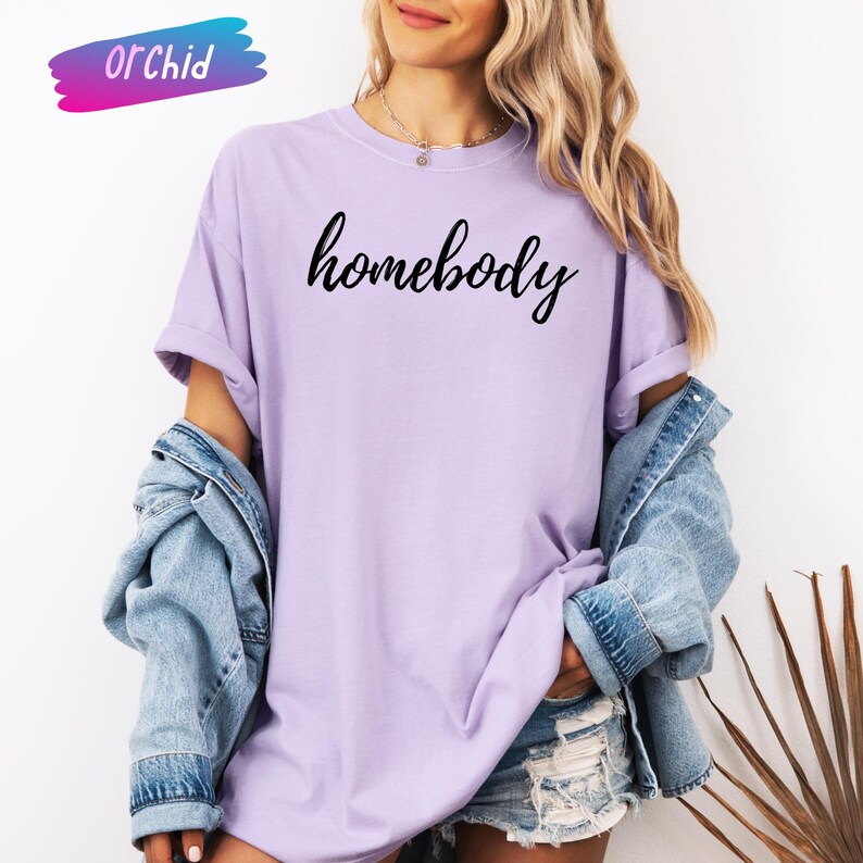 May include: A light purple t-shirt with the word "homebody" in black script. The shirt has rolled-up sleeves and is worn with a denim jacket. The word "Orchid" is written in a colorful font in the upper left corner.