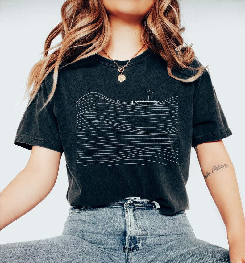 May include: Black t-shirt with a white line drawing of a boat on the ocean. The design features horizontal lines representing waves. The shirt is a dark charcoal color and has a vintage, worn look.
