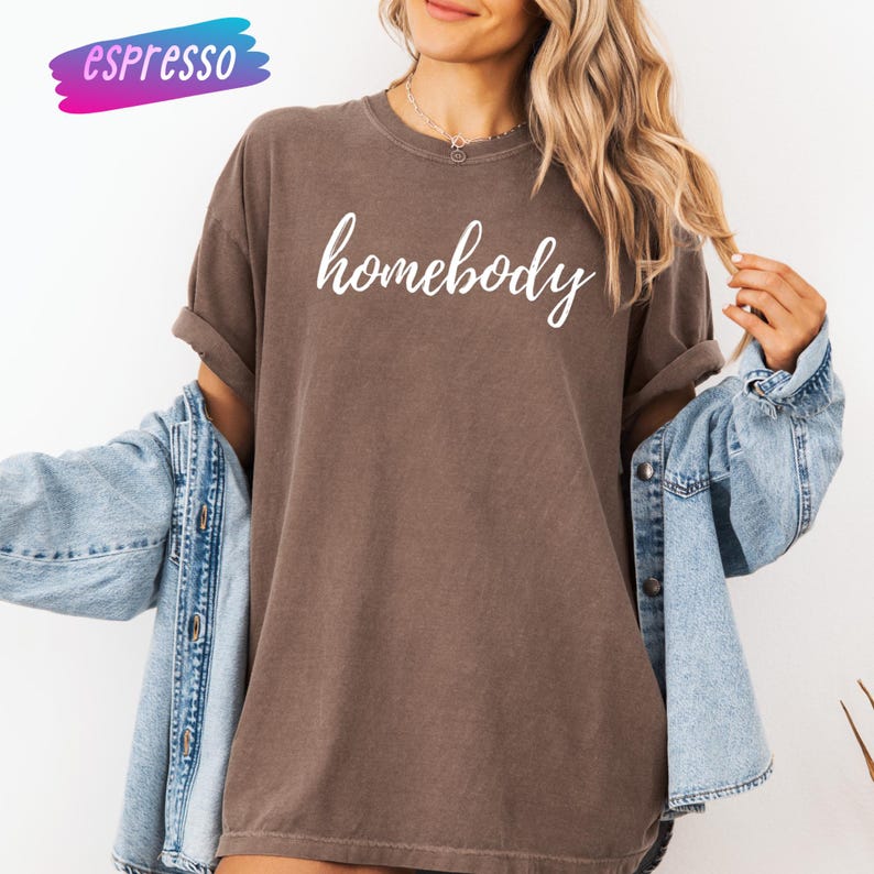 May include: A brown t-shirt with the word "homebody" in white script. The shirt is worn with a denim jacket. The word "espresso" is in a colorful banner in the upper left corner.