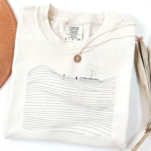 May include: Cream-colored Comfort Colors t-shirt with a black line drawing of a boat on wavy lines. A gold necklace with a pendant is draped on the shirt. The shirt has a tag that says "Comfort Colors".