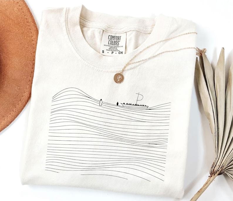 May include: Cream-colored Comfort Colors t-shirt with a black line drawing of a boat on wavy lines. A gold necklace with a pendant is draped on the shirt. The shirt has a tag that says "Comfort Colors".
