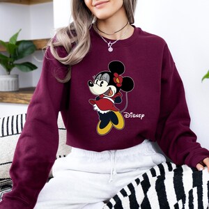 May include: A maroon cropped sweatshirt with a cartoon image of Minnie Mouse in a red dress and yellow shoes. The text "Disney" is printed on the bottom right corner of the sweatshirt.