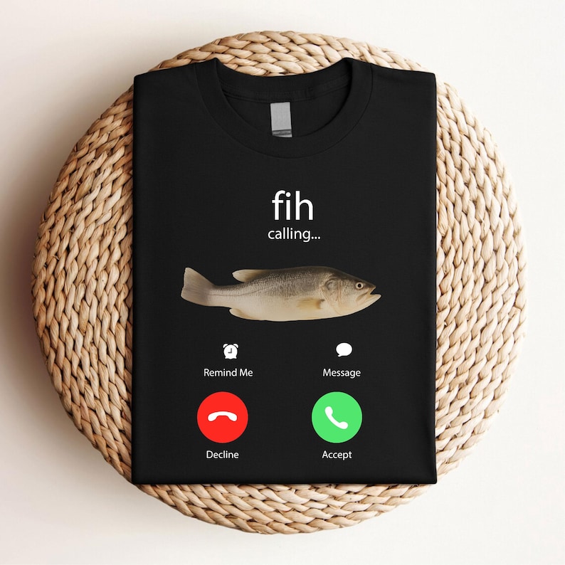 May include: Black t-shirt featuring a fish graphic with the text "fih calling...". Below the fish are icons for "Remind Me" and "Message", with "Decline" and "Accept" buttons. The shirt is displayed on a woven surface.