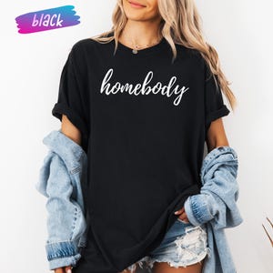 May include: Black t-shirt with the word "homebody" in white script. The shirt is worn with a denim jacket and shorts. A colorful "black" label is in the upper left corner.