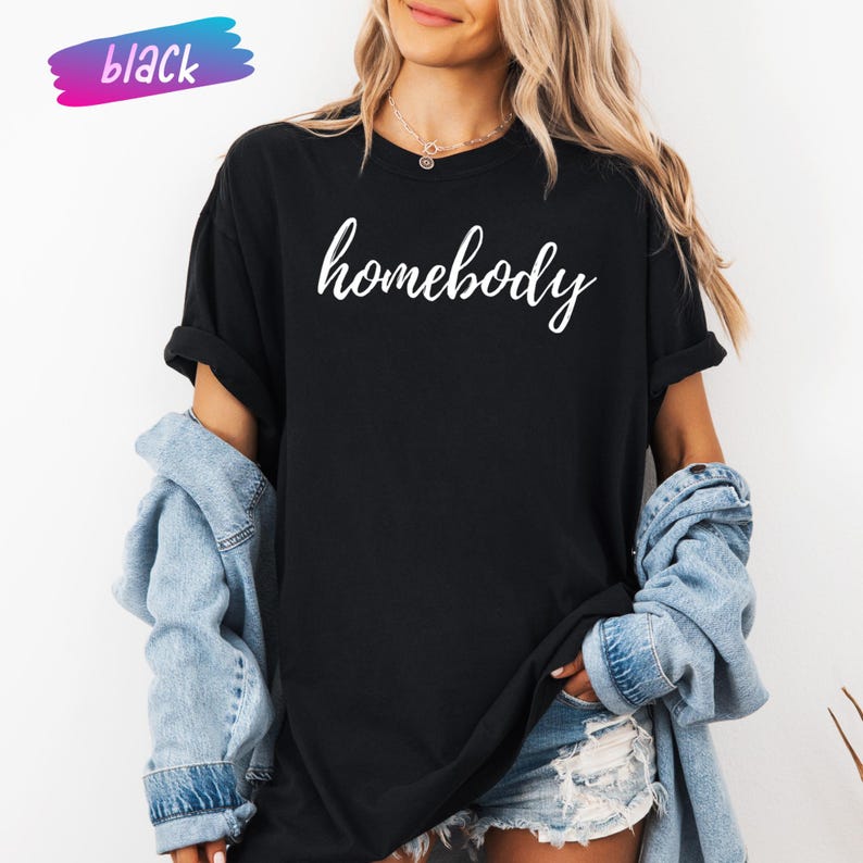 May include: Black t-shirt with the word "homebody" in white script. The shirt is worn with a denim jacket and shorts. A colorful "black" label is in the upper left corner.