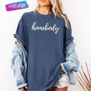May include: A navy blue t-shirt with the word "homebody" in white script. The shirt is worn with a light blue denim jacket. The word "bluejean" is written in a pink and blue font in the upper left corner.