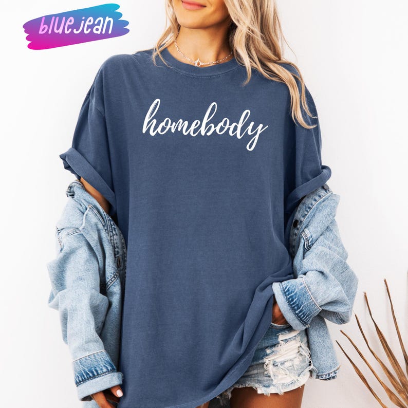 May include: A navy blue t-shirt with the word "homebody" in white script. The shirt is worn with a light blue denim jacket. The word "bluejean" is written in a pink and blue font in the upper left corner.