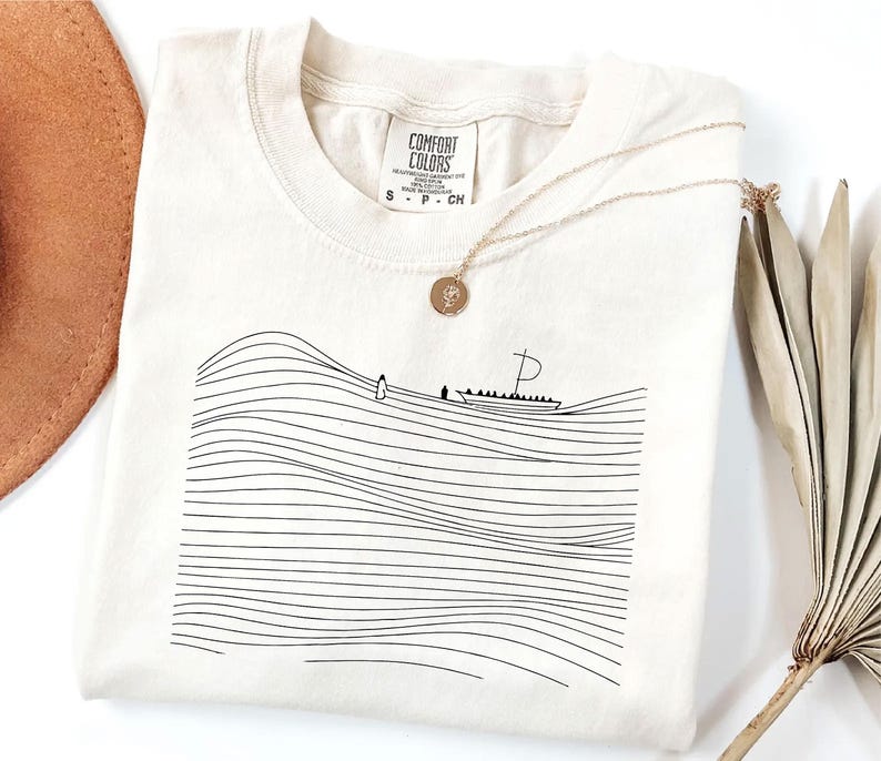 May include: Cream-colored Comfort Colors t-shirt with a black line drawing of a boat on wavy lines. A gold necklace with a pendant is draped on the shirt. The shirt has a tag that says "Comfort Colors".