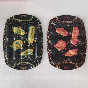 Op de afbeelding: Twee zwarte Tefal bakvormen met cartoon voedselvormen. Eén vorm heeft gele en groene ontwerpen, de andere heeft roze en gele ontwerpen. De vormen hebben de tekst "Tefal Made in France".