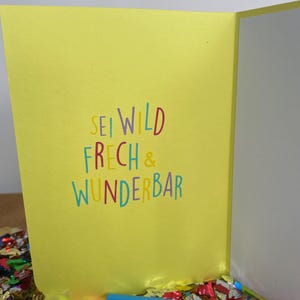 Puede incluir: Tarjeta amarilla con el texto "Sei wild, frech & wunderbar" en letras de colores. La tarjeta est&aacute; sobre una superficie cubierta de confeti de colores.