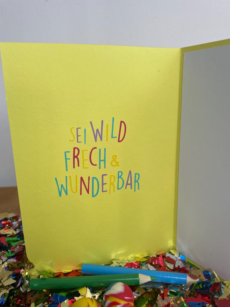 Puede incluir: Tarjeta amarilla con el texto "Sei wild, frech & wunderbar" en letras de colores. La tarjeta est&aacute; sobre una superficie cubierta de confeti de colores.