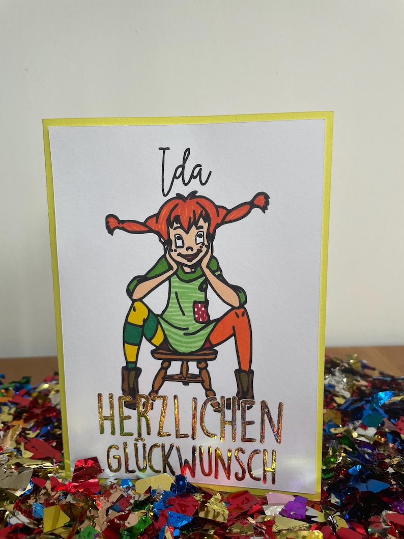 Puede incluir: Una tarjeta amarilla con una ilustraci&oacute;n de dibujos animados de una ni&ntilde;a con cabello rojo y un vestido verde. El texto "Ida" est&aacute; por encima de la ilustraci&oacute;n y "Herzlichen Gl&uuml;ckwunsch" est&aacute; por debajo de la ilustraci&oacute;n.