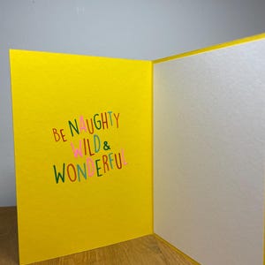 Puede incluir: Una tarjeta amarilla con el texto "Be Naughty Wild & Wonderful" en letras de colores.