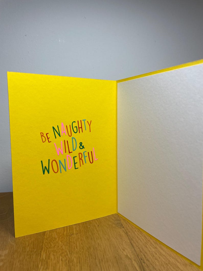 Puede incluir: Una tarjeta amarilla con el texto "Be Naughty Wild & Wonderful" en letras de colores.