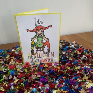 Puede incluir: Una tarjeta amarilla con una ilustraci&oacute;n de dibujos animados de una ni&ntilde;a con cabello rojo y una camisa verde. El texto "Ida" est&aacute; escrito en la parte superior de la tarjeta. El texto "Herzlichen Gl&uuml;ckwunsch" est&aacute; escrito en la parte inferior de la tarjeta.