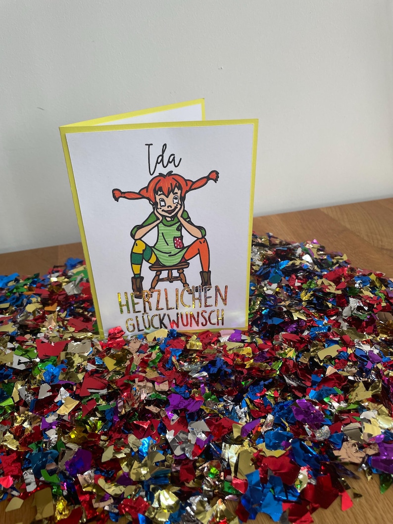 Puede incluir: Una tarjeta amarilla con una ilustraci&oacute;n de dibujos animados de una ni&ntilde;a con cabello rojo y una camisa verde. El texto "Ida" est&aacute; escrito en la parte superior de la tarjeta. El texto "Herzlichen Gl&uuml;ckwunsch" est&aacute; escrito en la parte inferior de la tarjeta.