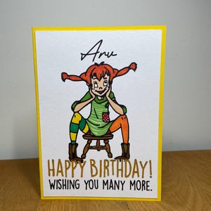 Puede incluir: Una tarjeta amarilla con una ilustraci&oacute;n de dibujos animados de una ni&ntilde;a con cabello rojo y un vestido verde. El texto "Happy Birthday! Wishing you many more." est&aacute; escrito en negro en la tarjeta.