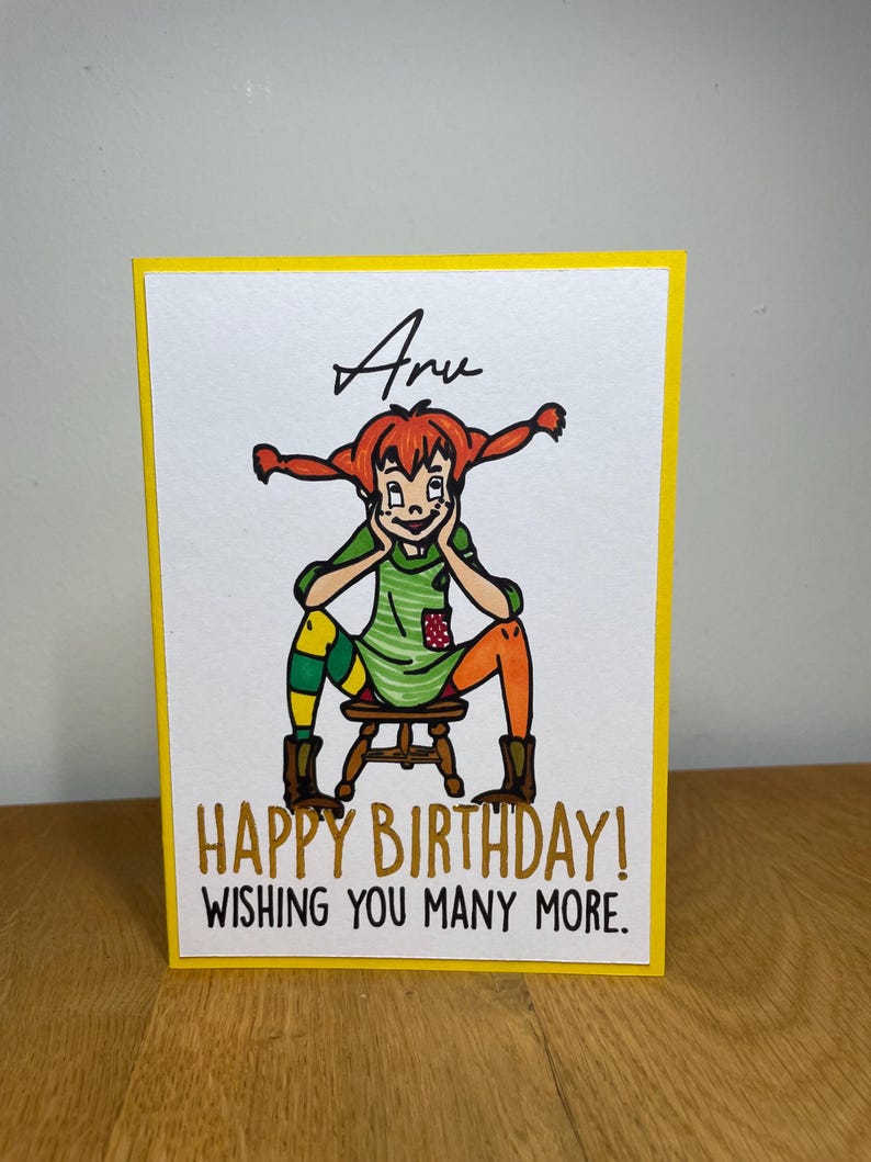 Puede incluir: Una tarjeta amarilla con una ilustraci&oacute;n de dibujos animados de una ni&ntilde;a con cabello rojo y un vestido verde. El texto "Happy Birthday! Wishing you many more." est&aacute; escrito en negro en la tarjeta.