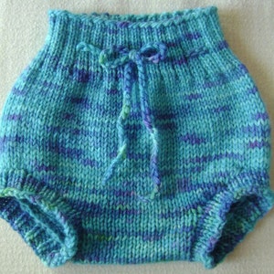 May include: Hand-knitted baby bloomers in shades of turquoise, blue, and purple. The bloomers feature a drawstring waist and a textured knit pattern. The garment is designed to be worn by a baby.