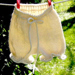 May include: Cream-colored knitted baby shorts with a drawstring waist and ruffled edges, hanging on a clothesline with a red clothespin. The shorts are made of soft yarn and appear handmade, suggesting a cozy and delicate design.