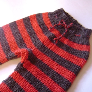 May include: Hand-knitted striped pants in red and dark gray. The pants feature a drawstring at the waist and are made of a soft, textured yarn. The stripes run horizontally across the legs.