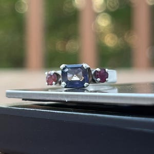 May include: A silver ring with a large, rectangular, dark blue gemstone in the center, flanked by two smaller, round, red gemstones. The ring is set on a black surface with a blurred green and brown background.