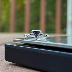 May include: A silver ring with a triangular, purple gemstone. The ring's band features a delicate, openwork design with small circular accents. The ring is set against a blurred background of green foliage.