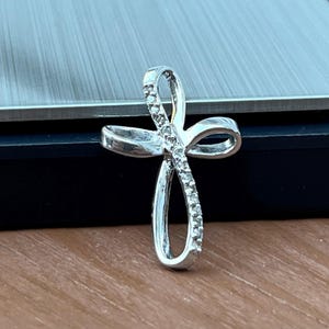 May include: A silver-colored pendant in the shape of a cross. The cross features a looped design with a row of small, clear stones along one side. The pendant is displayed on a wooden surface.