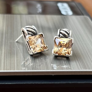 May include: A pair of silver stud earrings with square, light orange gemstones. The earrings have a textured design above the gemstone. The earrings are displayed on a silver surface.