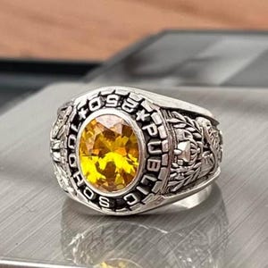 May include: A silver-toned class ring with a large, oval, yellow gemstone at its center. The ring is engraved with the words "SCHOOL PUBLIC" and "1959" around the gemstone. Intricate details and designs are visible on the ring's sides.