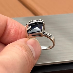 May include: A silver ring featuring a large, rectangular, dark blue gemstone. The ring has a decorative band with small, embedded stones around the gemstone and on the sides of the band. The ring is held by a person's hand.