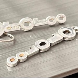 May include: Silver-toned earrings with a key-like design. Each earring features a series of circular and rectangular elements. One earring has a hook closure, while the other has a post. The earrings are displayed on a textured, metallic surface.