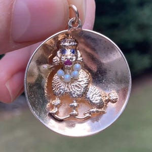 May include: A gold charm pendant featuring a poodle with blue eyes, a red ruby nose, and a pearl necklace. The poodle is standing on its hind legs and has its front paws raised.