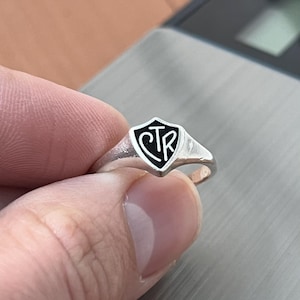 May include: A silver signet ring with a shield-shaped face. The shield has a black enamel background with the letters "CTR" in white. The ring is held up by a hand.