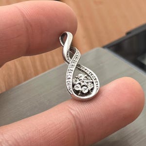 May include: A silver pendant in an infinity symbol design, encrusted with small, sparkling gemstones. The pendant features a cluster of gemstones at the bottom, adding a touch of elegance. The jewelry is held between fingers, showcasing its size and detail.