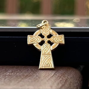 May include: A gold-colored Celtic cross pendant with intricate knotwork design. The cross features a circular halo and a detailed pattern on each arm. The pendant is displayed on a black surface.