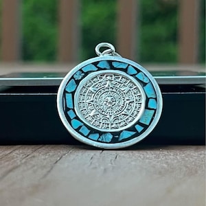 May include: A circular silver pendant featuring a central embossed Aztec sun calendar design surrounded by a border of inlaid light blue turquoise stone fragments. The pendant includes a small silver bail at the top for attachment to a chain.