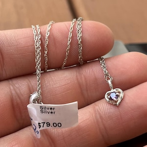 May include: A silver necklace with a heart-shaped pendant. The pendant features a small, purple gemstone set within a delicate silver frame. The necklace is displayed with a price tag that reads "Silver" and "$79.00".