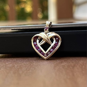 May include: A gold-toned heart pendant with a ruby gemstone border and a small ruby accent at the top. The pendant has an intertwined design and is set against a blurred background.