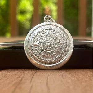May include: A circular silver pendant featuring a detailed embossed design of a sun face at the center, surrounded by intricate geometric patterns and symbols. The pendant has a polished metallic finish and a small loop at the top for attachment.