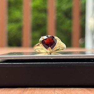 May include: A gold ring featuring a heart-shaped, deep red gemstone. The ring's band is a polished gold, and the gemstone is set in a prong setting. The ring is displayed on a black surface with a blurred green background.