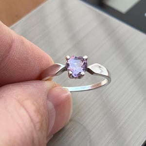 May include: A silver ring featuring a heart-shaped, light purple gemstone. The ring's band is a simple, polished silver, with the gemstone set in a four-prong setting. The ring is held against a gray surface.