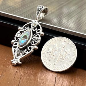 May include: A silver pendant with an oval abalone shell center, showcasing iridescent blues and greens. The pendant has an ornate, filigree design with small, decorative accents. A US dime is next to the pendant for size comparison.