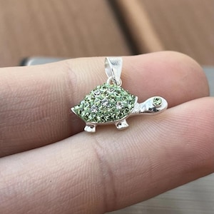 May include: A silver-toned turtle pendant with a green, gem-studded shell and a single green gem eye. The pendant is small and detailed, with a loop for a chain.